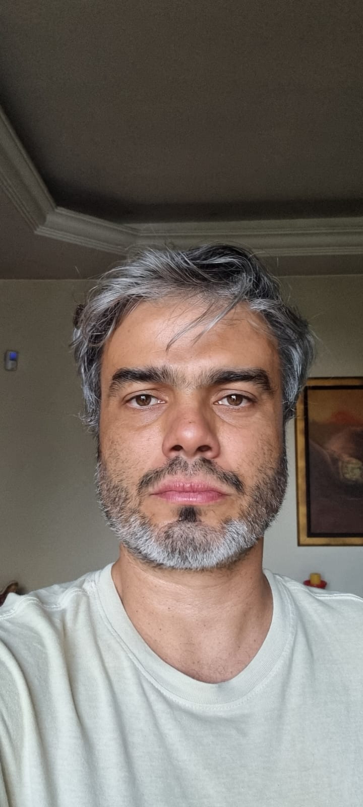 Homem branco de cabelos curtos e grisalhos, sorri com barba também grisalha, veste uma camiseta branca. Atrás um fundo preto.