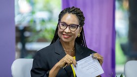 Magali Dantas, uma mulher negra, de cabelos longos trançados e presos no topo para trás. Ela sorri, tem olhos escuros, usa óculos de armações escuras, brincos dourados e batom vermelho.