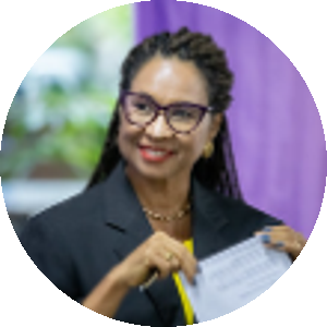Magali Dantas, uma mulher negra, de cabelos longos trançados e presos no topo para trás. Ela sorri, tem olhos escuros, usa óculos de armações escuras, brincos dourados e batom vermelho.