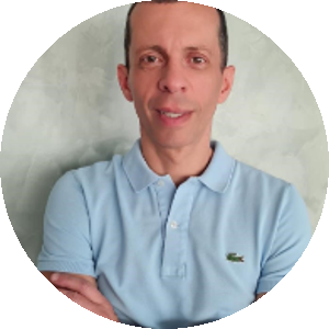 Homem branco de cabelos curtos pretos. Veste uma camiseta polo azul clara. Está em pé, com um leve sorriso no rosto e com os braços cruzados. Ao fundo, uma parede em tom esverdeado.