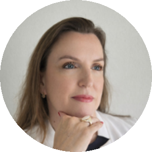 mulher branca, cabelos lisos e castanhos. Olhos azuis. Usa um blazer branco e uma blusa preta. Está com a mão apoiando o queixo.