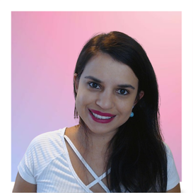 mulher de cabelo castanho longo penteado de lado. Veste uma blusa branca. Está com um sorriso largo, com um fundo rosa.