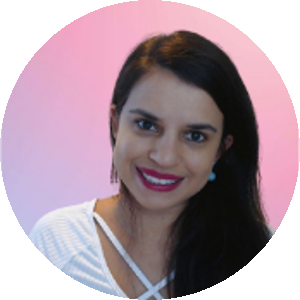 mulher de cabelo castanho longo penteado de lado. Veste uma blusa branca. Está com um sorriso largo, com um fundo rosa.