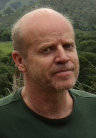 Homem branco, com olhar sério para a câmera. Olhos claros e cabelos raspados. Veste uma blusa verde.