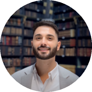 Marlon Amorim, homem branco, de cabelos e olhos escuros, barba baixa e desenhada. Ele sorri e ao fundo há estantes com livros.