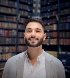 Marlon Amorim, homem branco, de cabelos e olhos escuros, barba baixa e desenhada. Ele sorri e ao fundo há estantes com livros.