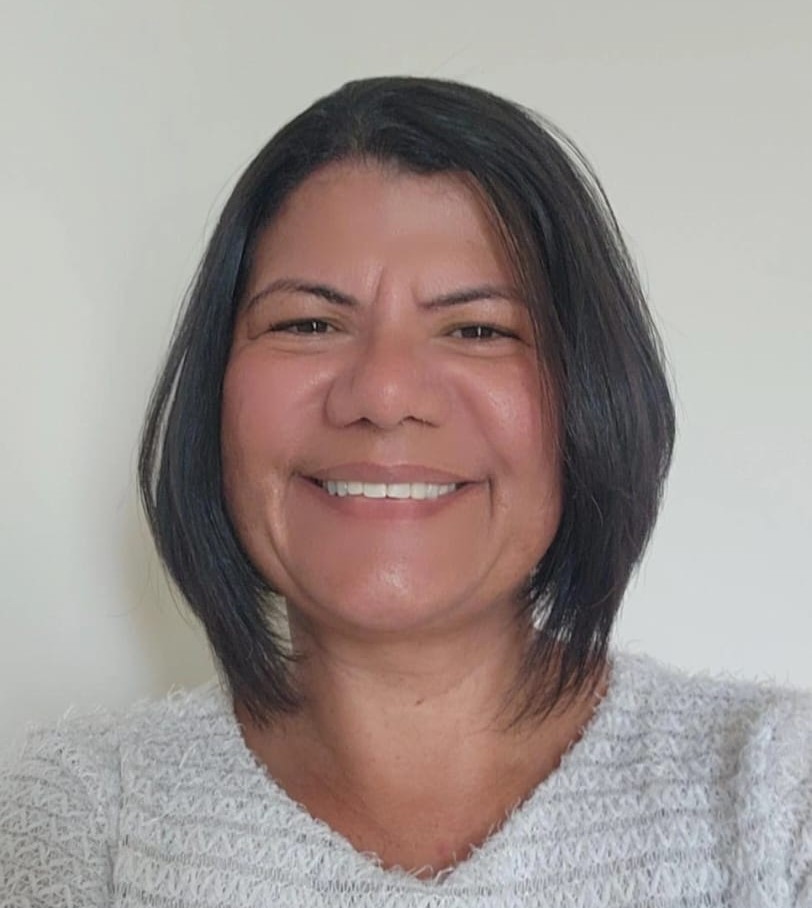 Mulher de cabelo curto e liso, sorrindo em frente a um fundo claro. Ela veste uma blusa clara de textura felpuda e transmite uma expressão acolhedora e simpática.