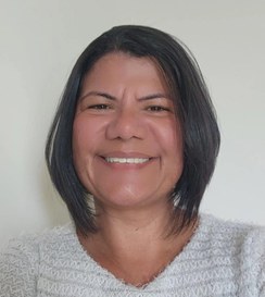 Mulher de cabelo curto e liso, sorrindo em frente a um fundo claro. Ela veste uma blusa clara de textura felpuda e transmite uma expressão acolhedora e simpática.