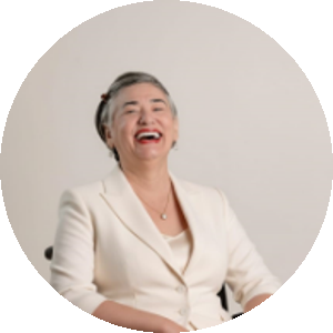 Mulher sentada em uma cadeira de rodas, sorrindo amplamente. Ela usa um terninho branco e brincos discretos, com o cabelo curto penteado para o lado. O fundo é neutro e claro, destacando sua expressão alegre e confiante.