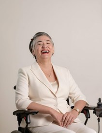 Mulher sentada em uma cadeira de rodas, sorrindo amplamente. Ela usa um terninho branco e brincos discretos, com o cabelo curto penteado para o lado. O fundo é neutro e claro, destacando sua expressão alegre e confiante.