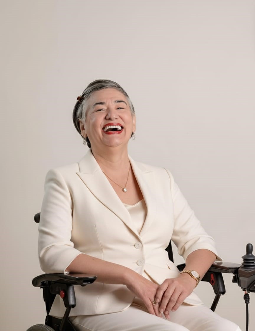 Mulher sentada em uma cadeira de rodas, sorrindo amplamente. Ela usa um terninho branco e brincos discretos, com o cabelo curto penteado para o lado. O fundo é neutro e claro, destacando sua expressão alegre e confiante.