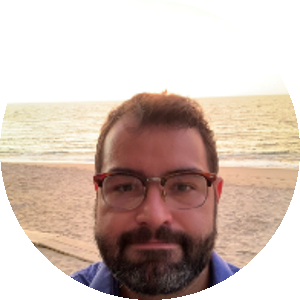 homem branco, cabelo e olhos castanhos. Usa óculos, barba e bigode. Usa uma camisa de botão azul e ao seu fundo uma praia.