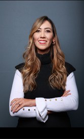 Mulher de cabelos longos e ondulados, com mechas loiras. Ela está de braços cruzados, sorrindo, vestindo uma blusa branca com colete preto e botões dourados, diante de um fundo cinza.