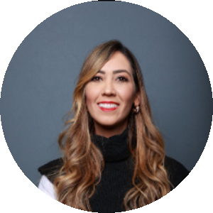 Mulher de cabelos longos e ondulados, com mechas loiras. Ela está de braços cruzados, sorrindo, vestindo uma blusa branca com colete preto e botões dourados, diante de um fundo cinza.