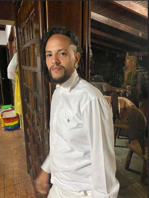 Lino Gabriel, um homem pardo, de olhos e cabelos escuros, cabelos cacheados e raspados na lateral esquerda, rosto ovalado e com bigode e cavanhaque. Ele usa brinco e uma camisa branca.