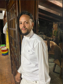 Lino Gabriel, um homem pardo, de olhos e cabelos escuros, cabelos cacheados e raspados na lateral esquerda, rosto ovalado e com bigode e cavanhaque. Ele usa brinco e uma camisa branca.