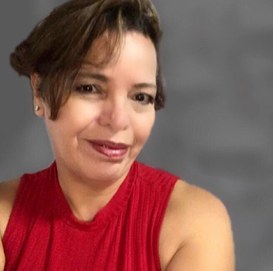 Mulher parda, usando uma blusa sem mangas na cor vermelha, cabelos curtos com uma franja lateral, brincos pequenos, batom vermelho e um sorriso discreto