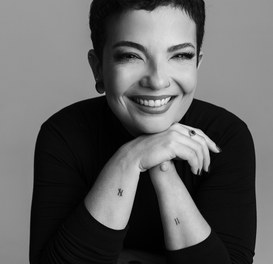 Fernanda Boaventura, mulher branca, de olhos castanhos e cabelos pretos bem curtos. Ela sorri com o rosto apoiado na mão direita e veste uma blusa preta de gola alta.