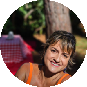 mulher branca de cabelos curtos e castanhos vestindo uma blusa de alça laranja. Está sentada e sorrindo. Ao seu fundo um gramado, uma árvore e uma mesa baixa com toalha quadriculada em vermelho e branco sobre um tapete vermelho.