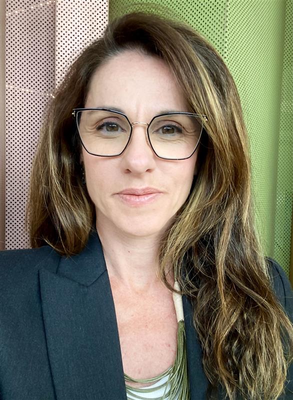 Claudia Wehbe, uma mulher branca, de cabelos castanhos claros e olhos castanhos. Ela usa óculos de armações pretas e finas e uma camisa verde escura.