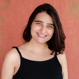 . Foto de Paloma Abelin, mulher branca de olhos verdes, cabelos lisos e escuros até o ombro. Ela veste uma blusa de alça preta e sorri.