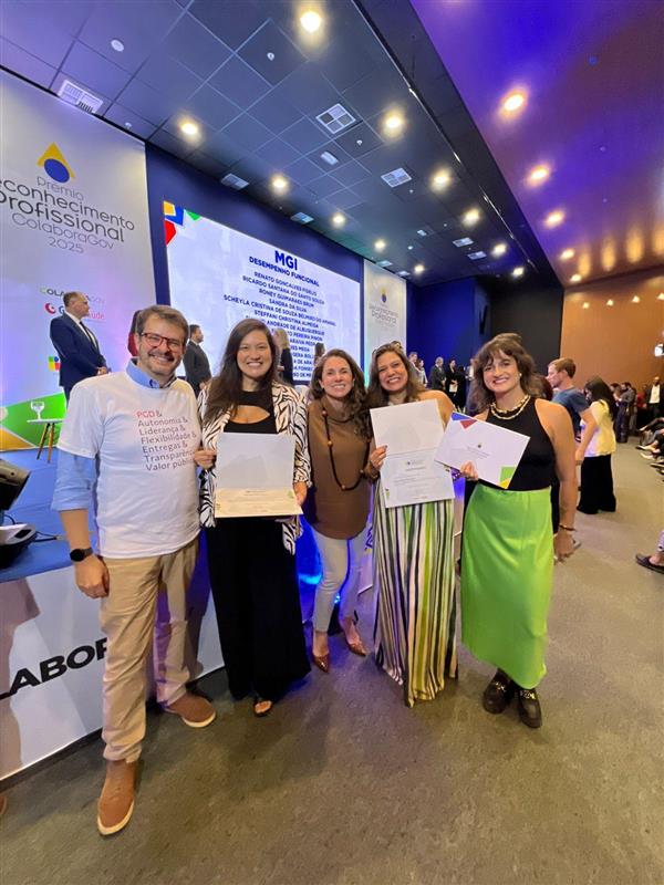 Grupo de seis pessoas em um palco de evento institucional, sorrindo e segurando certificados. Ao fundo, há um telão com identidade visual do MGI e do Prêmio de Reconhecimento Profissional 2025. As pessoas estão vestidas de forma social e casual, em clima de celebração, com outras pessoas e iluminação cênica ao redor.