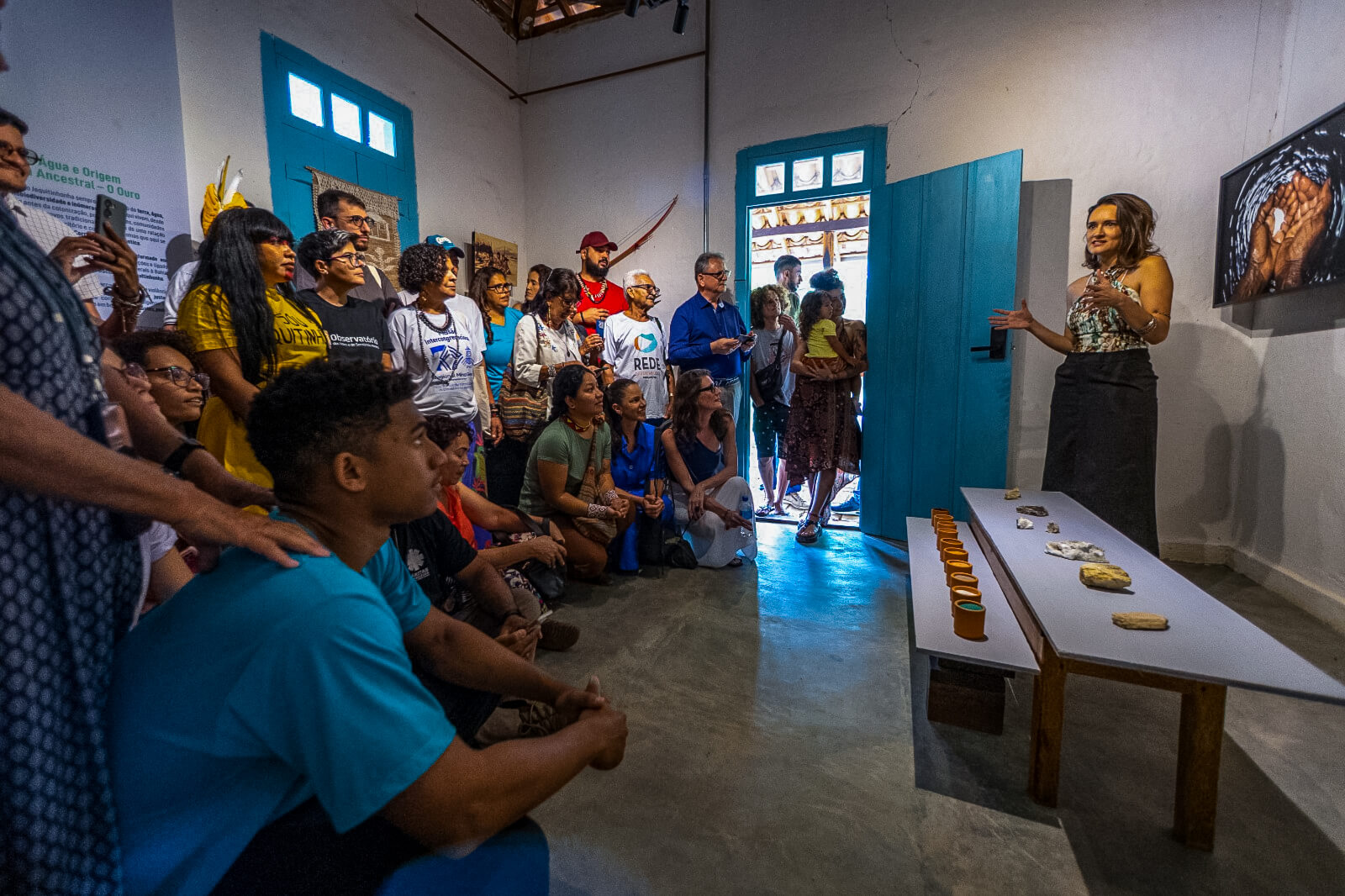 Foto de pessoas reunidas em um museu observando uma mulher falar.