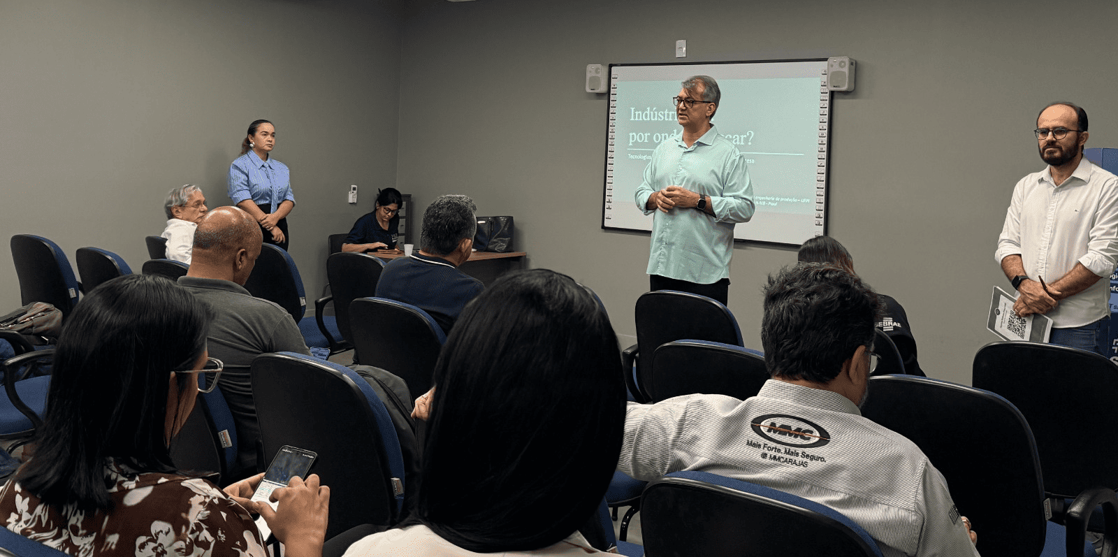 Sala de reuniões, com palestrantes à frente da plateia. O palestrante principal tem cabelo grisalhos e veste uma camisa verde clara com calça preta
