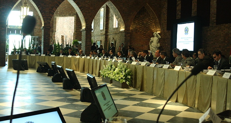 Imagem de uma mesa disposta em formato de L. Pessoas estão sentadas e promovem um debate.