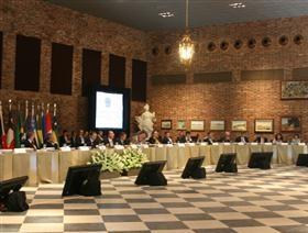 Plano geral de umas salão de cerimônias com várias pessoas sentadas atrás de uma bancada.