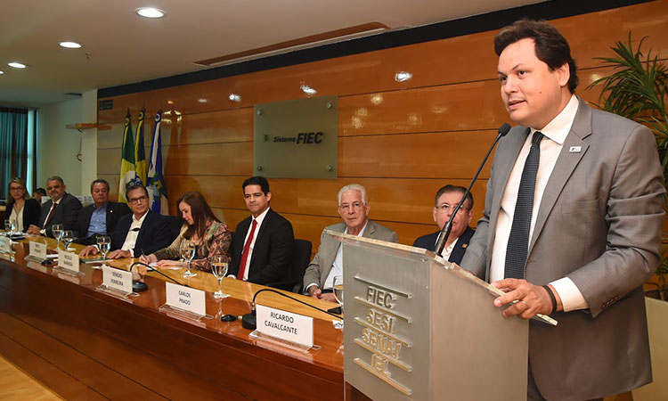 Apresentação do superintendente Marcelo Neves, durante evento na FIEC. A foto mostra o superintendente no púlpito e, ao lado, a mesa de abertura do evento, composta por oito pessoas.