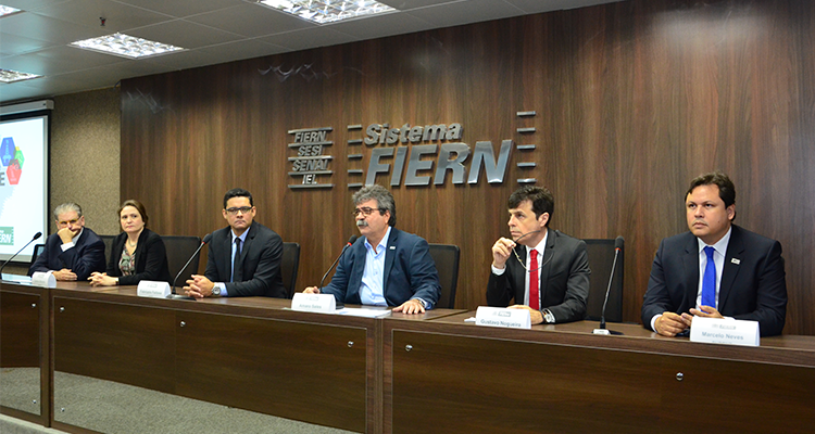 Mesa de abertura do evento composta por seis autoridades, entre elas o superintendente Marcelo Neves. Eles estão sentadoa lado a lado e atrás está escrito "Sistema FIERN".