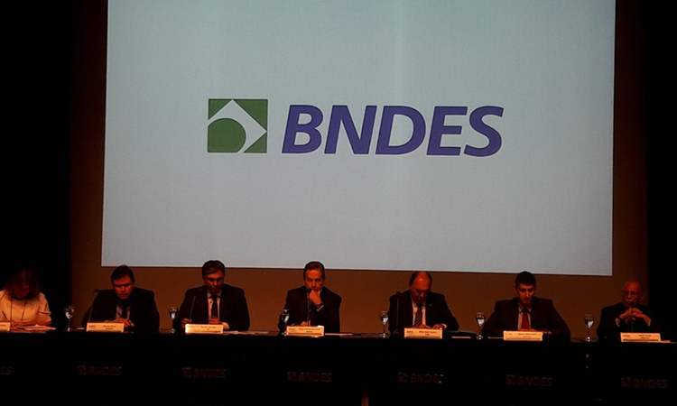 A foto mostra uma mesa com seis pessoas. Atrás delas, um telão exibe as letras "BNDES".