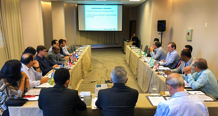 A foto traz pessoas sentadas em volta de um conjunto de mesas dispostas em formato da letra U. Há um telão no fundão no qual é projetada uma apresentação de slides. O público observa a tela e conversa.