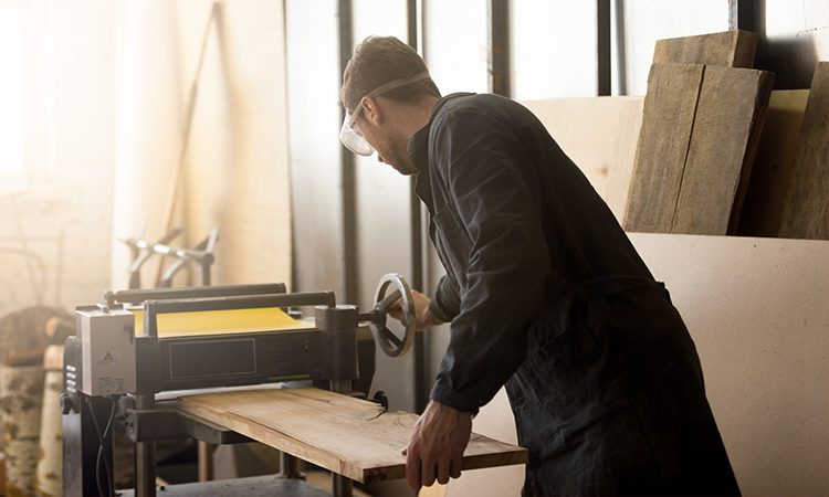 A imagem mostra um carpinteiro que lixa uma tábua de madeira em uma máquina. Ele está em sua própria marcenaria.