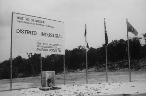 Foto em preto e branco que mostra a pedra fundamental do Distrito Industrial sobre um chão de barro, tendo logo acima uma placa com a inscrição "Ministério do Interior", "Superintendência da Zona Franca de Manaus", "Distrito Industrial" e "Aqui será implantado o desenvolvimento da Amazônia Ocidental". Ao lado da pedra estão três bandeiras (do Brasil, do Amazonas e da Suframa) e, ao fundo, a floresta.