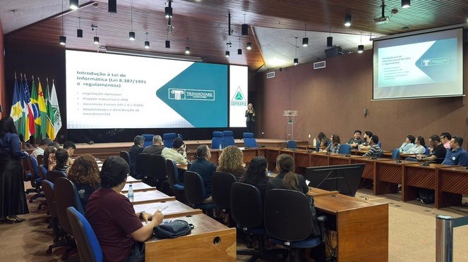 Foto mostra mulher falando em evento, observado por outras pessoas e com um telão ao fundo