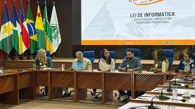 Foto mostra um homem falando em evento, observado por outras pessoas e com um telão ao fundo