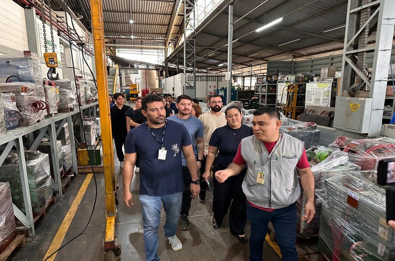 Grupo de alunos e representantes da empresa caminha por galpão industrial, observando máquinas e equipamentos durante visita técnica à fábrica