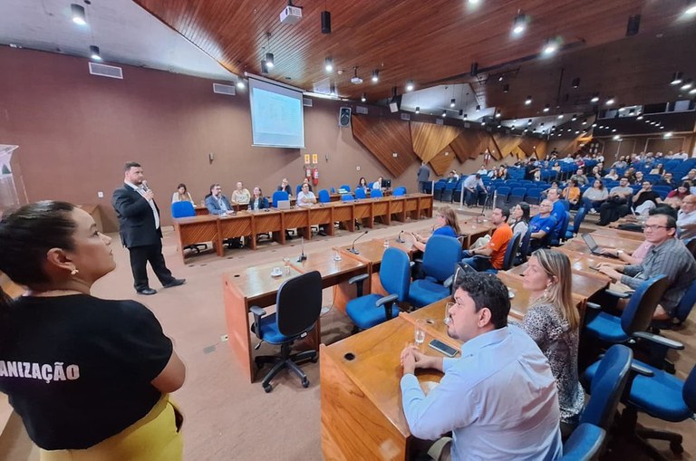 Foto mostra homem em pé falando ao microfone, diante de um auditório quase cheio de pessoas