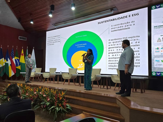 Foto mostra mulher falando em evento, com telão ao fundo