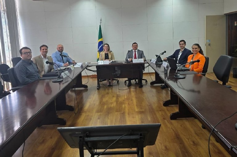 Foto mostra pessoas posando para fotografia durante reunião institucional, com um aparelho de televisão no primeiro plano e uma bandeira do Brasil ao fundo