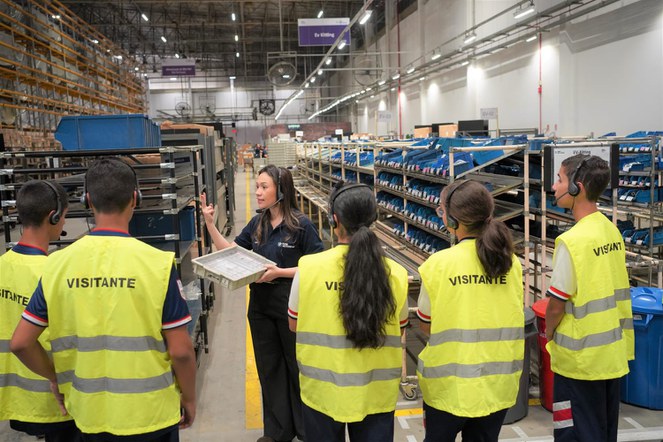 Foto mostra pessoas com jalecos amarelos recebendo uma explicação de uma mulher