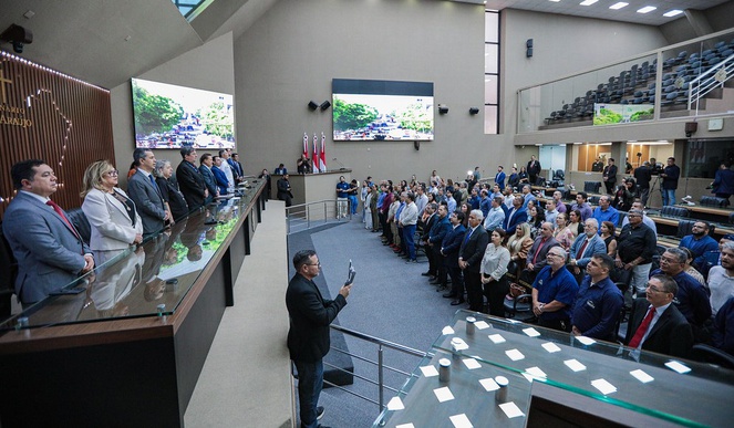 Foto mostra pessoas em pé dentro de um auditório com telões ao fundo