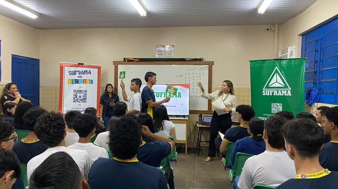 Foto mostra mulher de blusa bege clara, fazendo uma dinâmica com estudantes