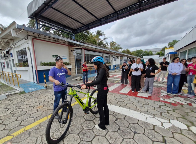 Foto mostra jovem de capacete e uma bicicleta, observada por instrutor e grupo de pessoas