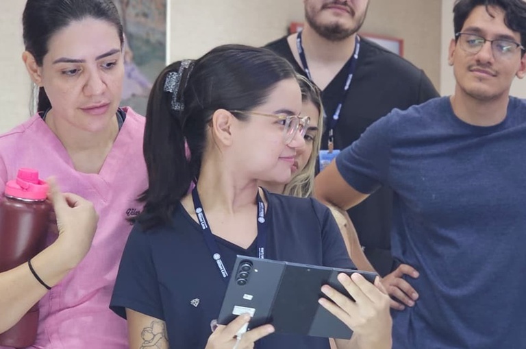 Foto mostra jovem de azul segurando um equipamento eletrônico, observada por outras pessoas