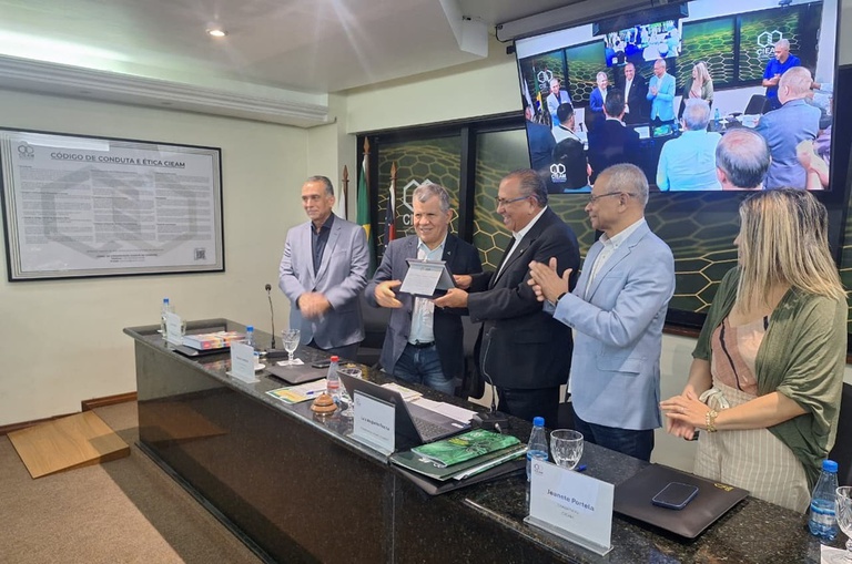 Foto mostra dois homens segurando uma placa de homenagem, observados por outras pessoas