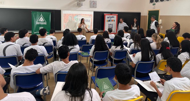 Foto mostra uma mulher falando, com baners atrás dela e pessoas observando