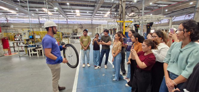 Foto mostra homem com capacete branco mostrando uma roda de bicicleta a um grupo de acadêmicos